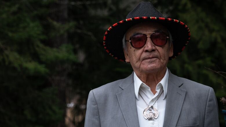 A man wearing a hat, sunglasses and a suit jacket stares seriously at the camera