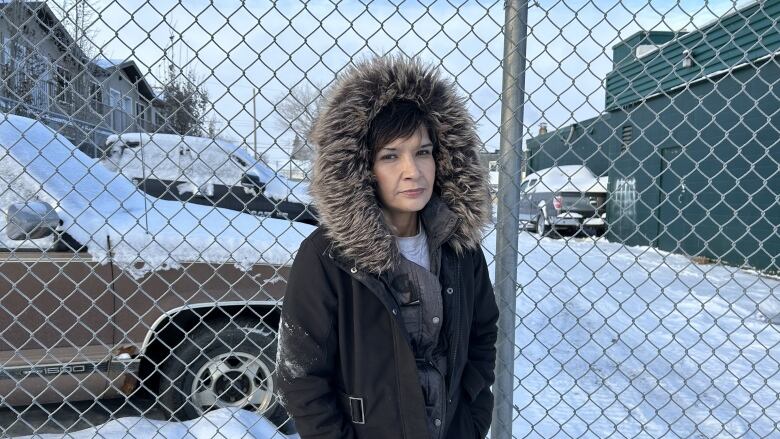 A middle-aged woman stands clad in a black parka against a wire mesh fence wall.