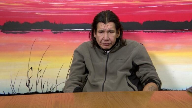 A middle-aged man sits at a table with a sunset landscape painted on the wall behind him.