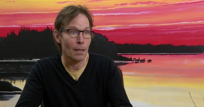 A middle-aged man sits at a table with a sunset landscape painted on the wall behind him.