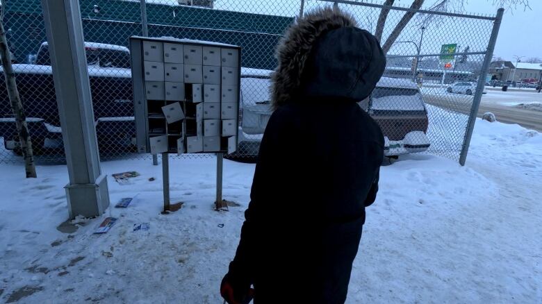 A woman dressed in parka looks at broken mail boxes in a unit. Her back is facing the camera.