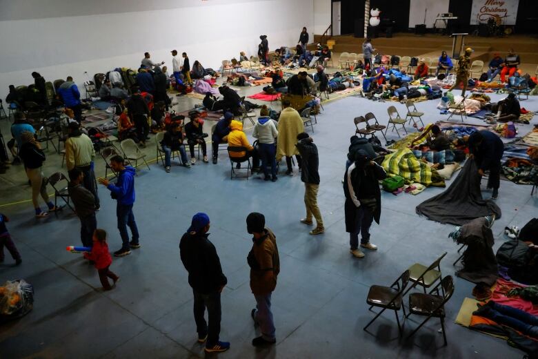 Dozens of people stand, sit on folding chairs, or lie under blankets on the floor of a large room.