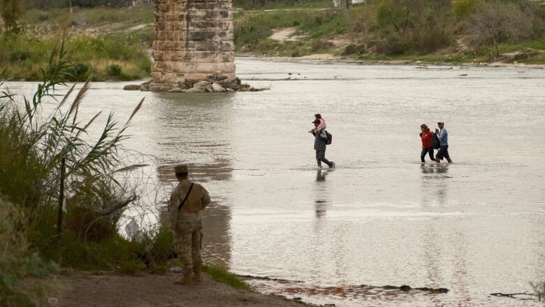 In the distance, three people, including one with a small child on their shoulders, walk across the shallow part of a river while a guard in a camouflage uniform watches them from the shore.