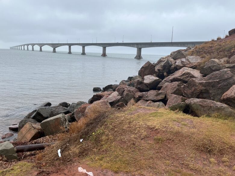 A large concrete bridge is seen in the Northumberland Straight, with red clay, green grass and large boulders in the foreground. 