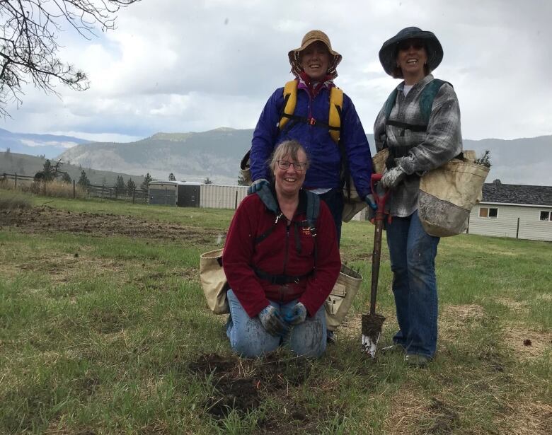 One woman kneels, and two women stand behind her, one with a shovel, on farmland, with a cloudy sky in the background.