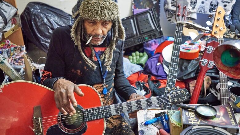 A man sits in a cluttered room full of guitars.