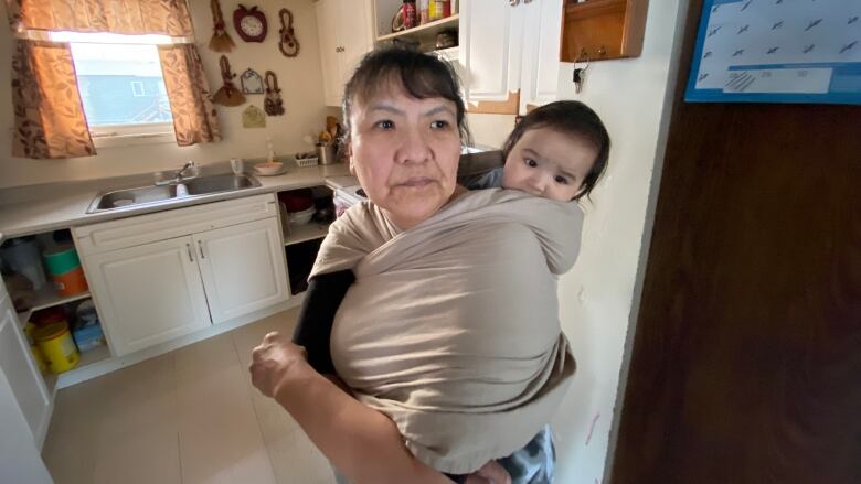 A woman stands in her kitchen with a baby wrapped around her back. 