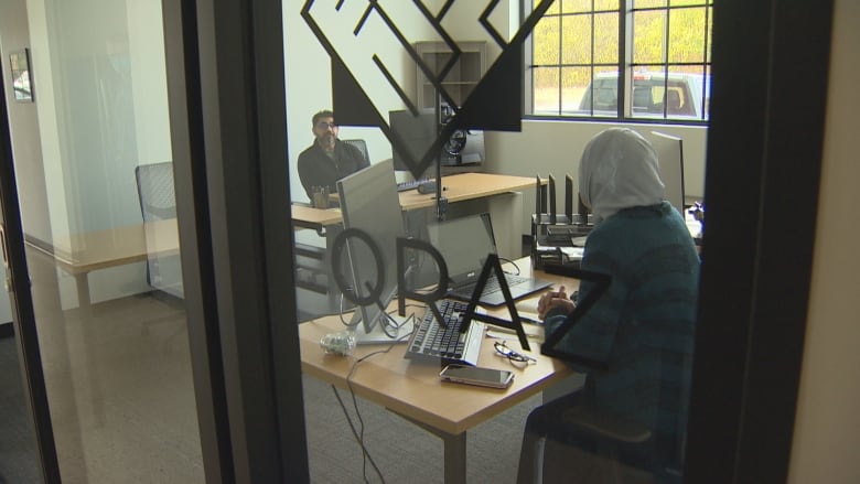 A man sits at a desk across from a woman, clad in headscarf; both are working at computers.