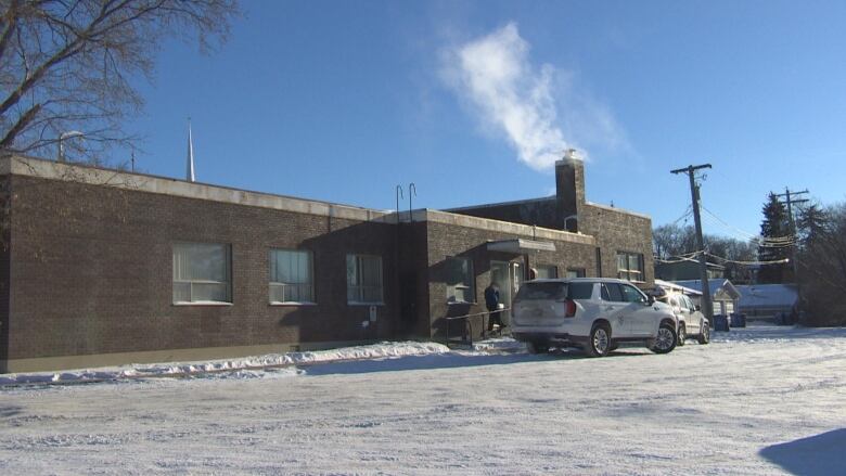 A large, single-storey brick building building is shown, with snow on the ground in front and vehicles parked by the door.