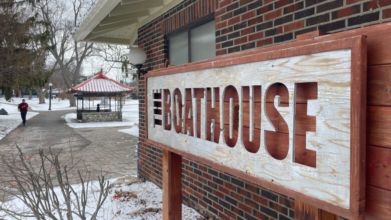 A wooden sign that says 'The Boathouse.' Snow is on the ground, a brick building on the right, a man jogging can be seen off to the left.