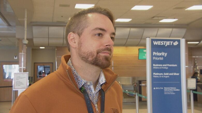 A man stands in an airport check-in area. His hair is parted to the left and he has a trimmed beard. He is wearing an orange collared fleece, a multi-coloured dress shirt and a lanyard.