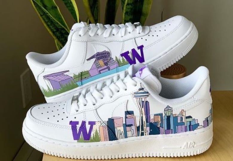 White sneakers are painted with an artistic version of the Seatle skyline in shades of blue, purple and pink on one side, and the University of Washington stadium on the other. They also have a large purple W on each side.