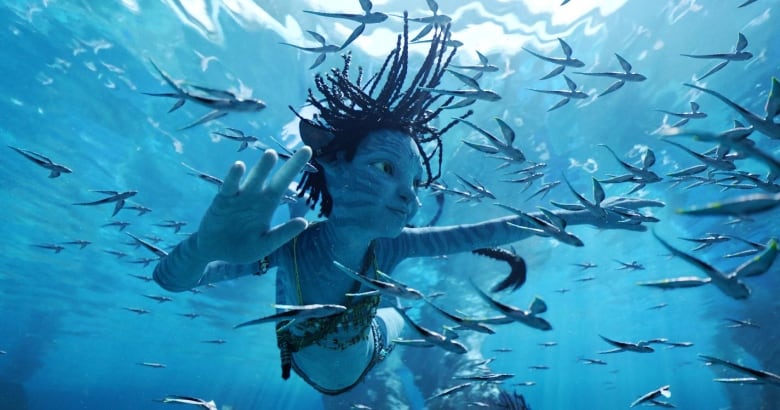 A humanoid blue alien with pointy ears and braided hair swims beneath the surface of the water surrounded by tiny fish-like creatures