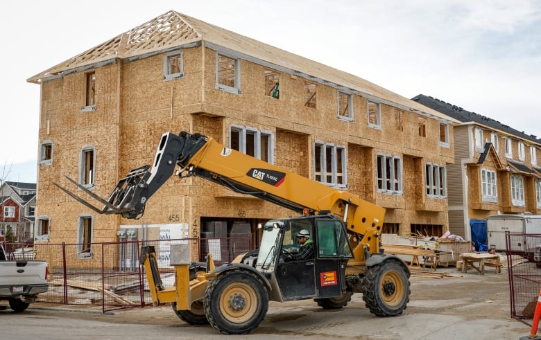 A yellow and black construction vehicle drives past an unfinished row of townhouses with light brown wooden frames and white window frames