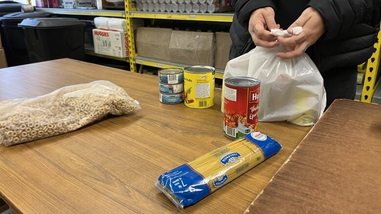 Cereal, tuna, canned fruit and pasta on a table, a pair of hands tying a bag closed.