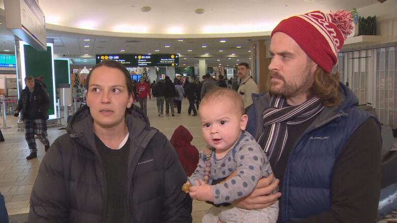 Two parents stand in the middle of an airport with a man in a tuque holding a baby.