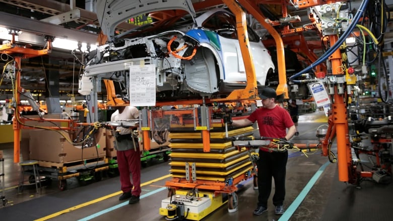 Two men stand either side of a partially-assembled car, which is raised into the air, inside a factory.