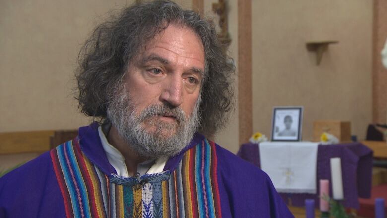 A priest wears a bright purple robe with multi-coloured Latin American stripes down the middle. Behind him, blurred, is a photo of Salomon Lzaro.