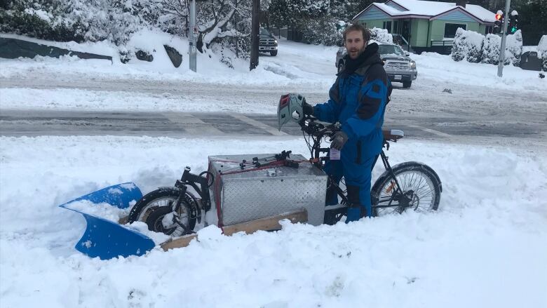 A man sits on a cargo bicycle with two shovels attached to the front using plywood. They are surrounded by heavy snow.