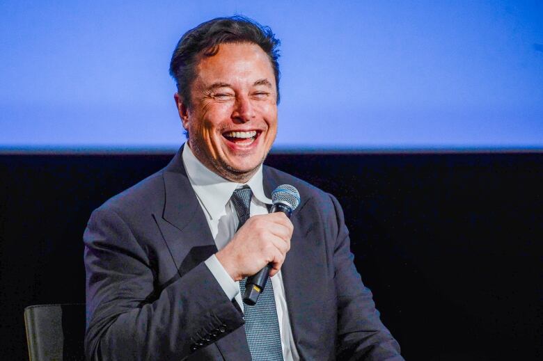 A seated man in a suit laughs while holding a microphone.