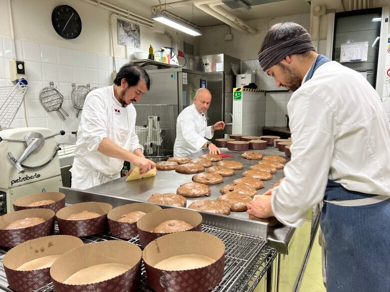 Three pastry chefs wearing white uniforms and aprons assemble panettone dough on steel table in bakery kitchen.