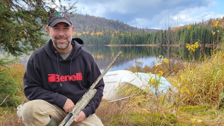 A man poses with a hunting rifle in front of a lake, while three dead birds lie at his feet.