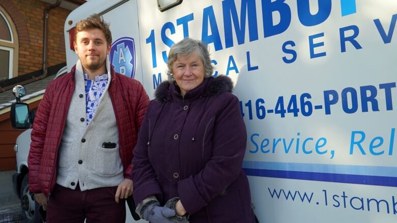 A young man and elderly woman wearing winter clothing stand outside in front of an ambulance.