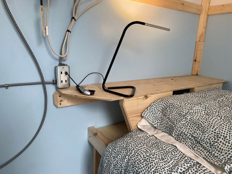 A lamp charged by a USB plug is on a shelf behind a bed.