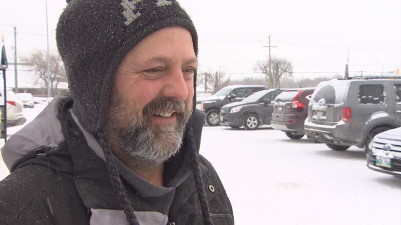 A bearded man wearing a grey toque smiles as he stands in a snowy parking lot.