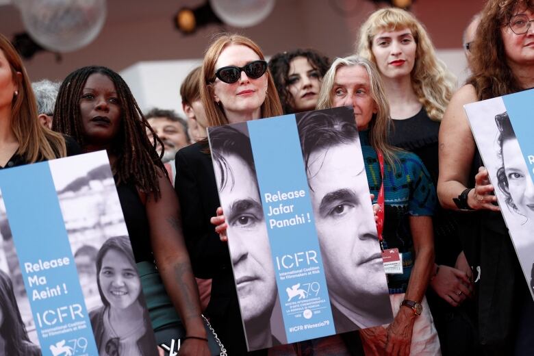 A woman wearing black sunglasses holds a blue poster with a black and white image of a man on it. Other people, holding similar posters, stand around her.