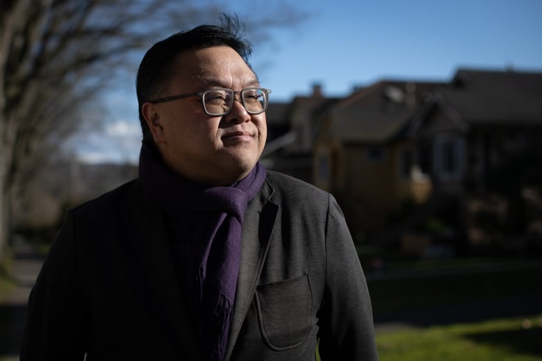 An Asian man wearing a black coat and a purple scarf stares off in the distance, near a row of single-family homes.