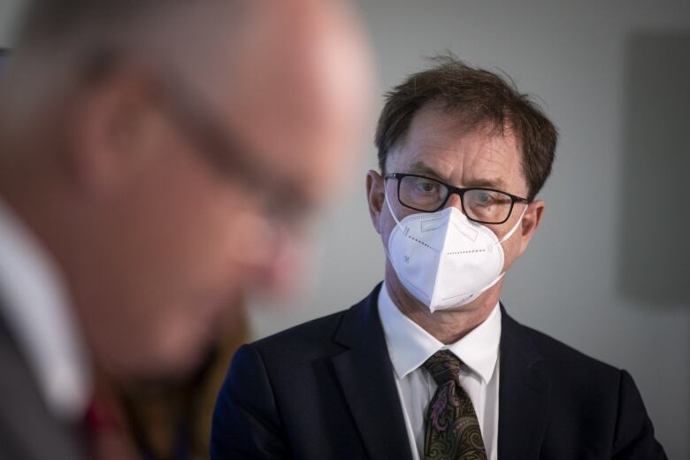 A man wears an N95 mask while standing in the background of a white man who is just out of focus.