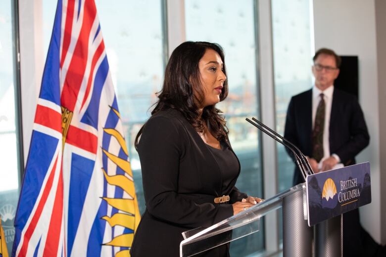 A woman speaks at a podium with the flag of British Columbia behind her. In the background, a man looks on.