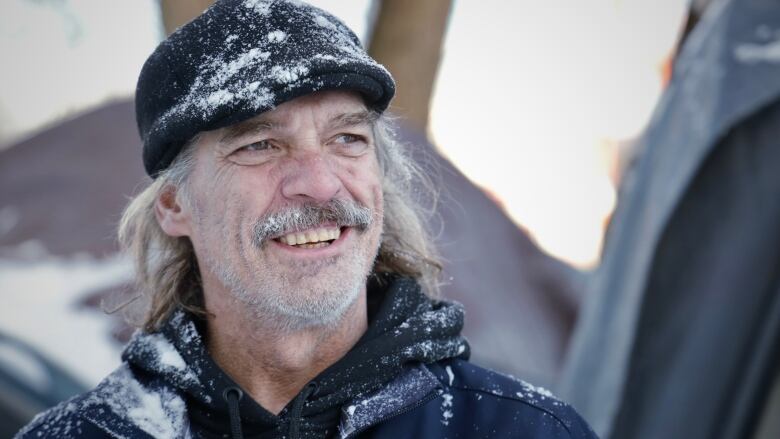 A man is pictured with a moustache and unshaven face, smiling. He's wearing a paperboy style hat and hooded sweatshirt, and snow is sticking to his clothes.