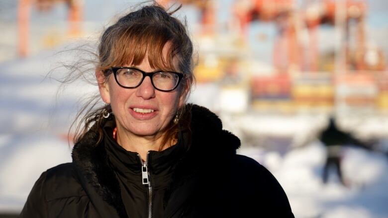 A woman wearing glasses is photographed looking directly at the camera. She has bangs, a winter jacket and the sun is shining on her face and the out-of-focus background.