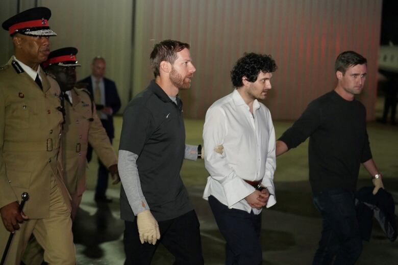 A man in a white dress shirt, in handcuffs, is escorted by two men. Police officers in khaki-coloured uniforms also flank the man.