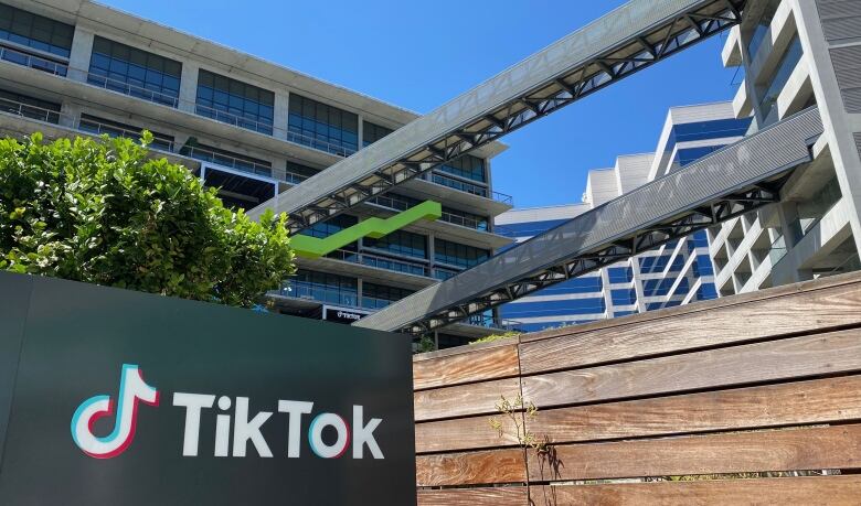 A black sign with white lettering next to a wooden fence, with a large office building in the background.