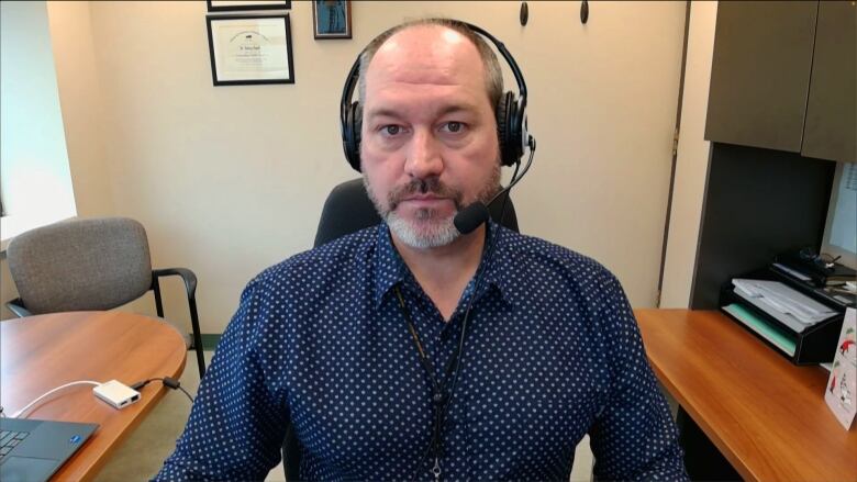 A man wearing a blue button-down shirt and headphones looks into the camera.