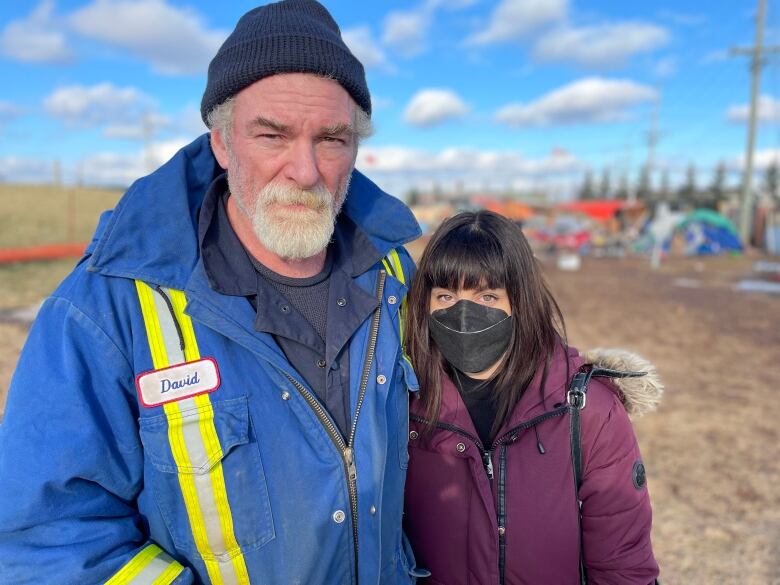 A man and woman stand next to each other dressed in winter gear, with a group of tents in the background behind them. 