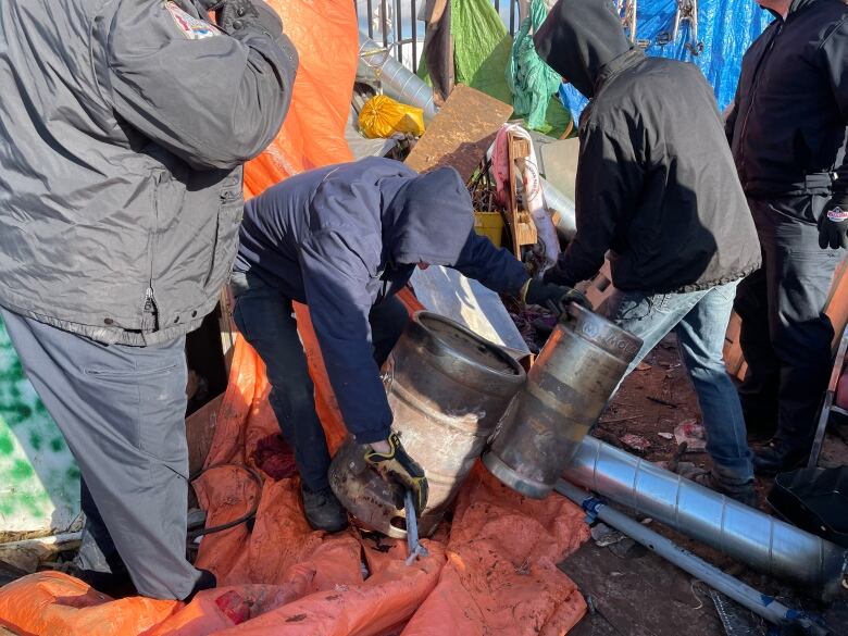 Several men wearing jackets with their hoods up lift metal kegs from the ground. 