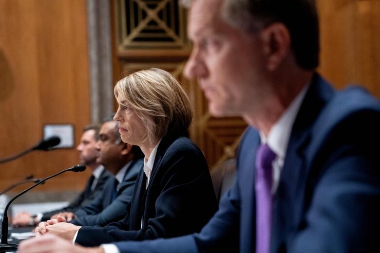 A blonde woman is pictured seated in a row between men wearing suits. They are all looking straight ahead, which is to the left of the image.