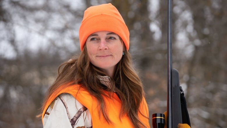 A woman wearing a camouflage jacket, bright orange vest and hat, is holding a hunting rifle. 