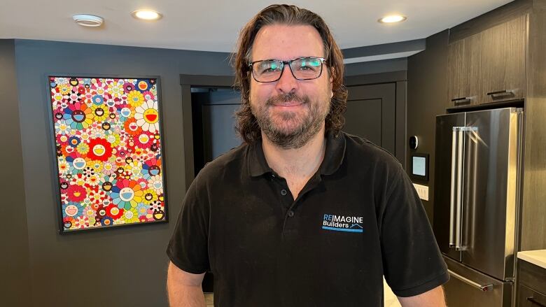 A man stands in his home, smiling, wearing a black polo t-shirt. There's a bright floral painting hanging in the background, on black walls.