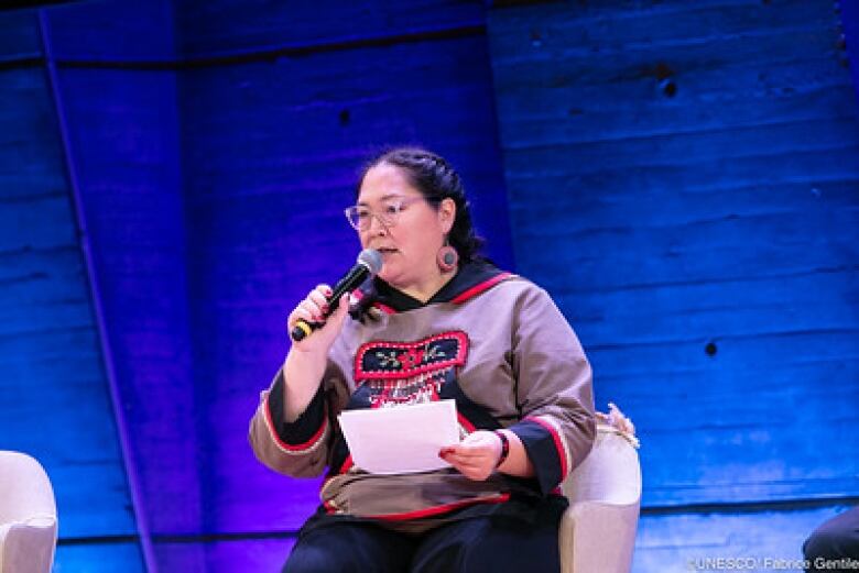 Aluki Kotierk sit on stage, speaking into a microphone. She wears beaded earrings and a matching beaded shirt. 