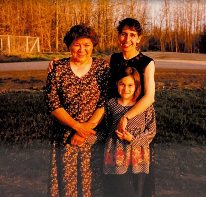 Three woman of different generations stand with their arms around each other smiling at the camera.