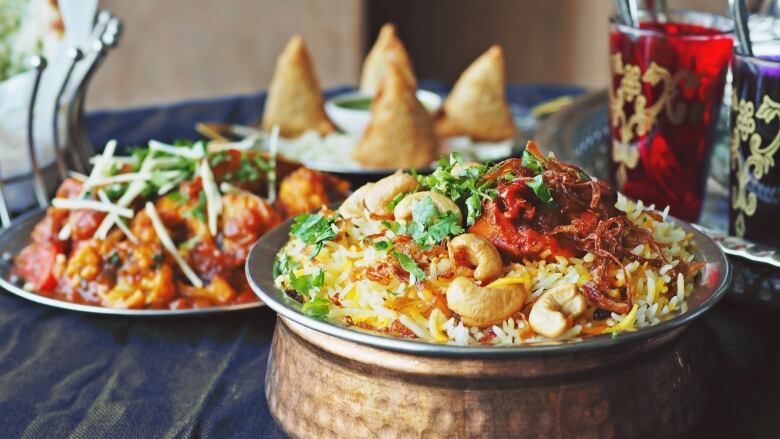 A bowl of biriyani, an Indian rice dish, sits in the foreground on a table. In the background, blurred visuals of curried potatoes and cauliflower, as well as triangular samosas are visible.