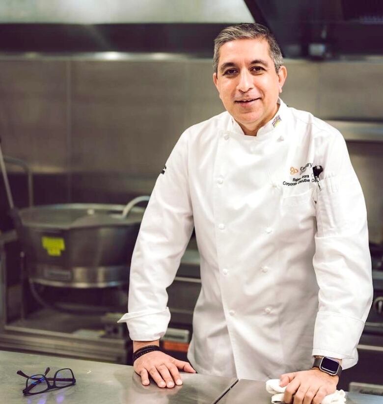 A South Asian man in a chef's uniform stands in a kitchen.