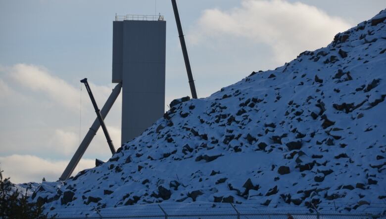 A mining headframe and cranes poke out from behind a big pile of rock, partly covered in snow. 