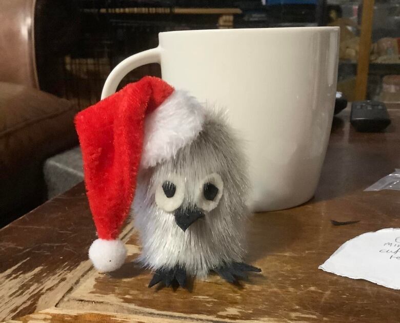 A hand-crafted owl made of fur wearing a santa hat. It sits next to a coffee mug.