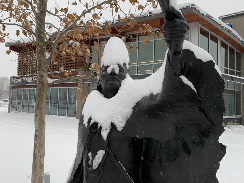 A statue of an Indigenous chief raising a feather is covered with snow, with a building in the background. 